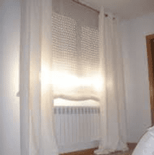 Ventana luminosa con cortinas transparentes y persianas cerradas sobre un radiador blanco en una habitación de color claro.