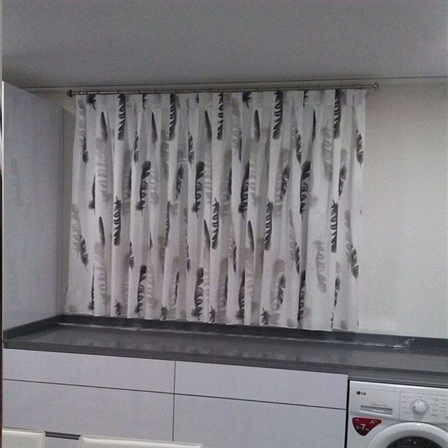 Cocina pequeña con una cortina blanca estampada sobre la encimera y una lavadora a la derecha.