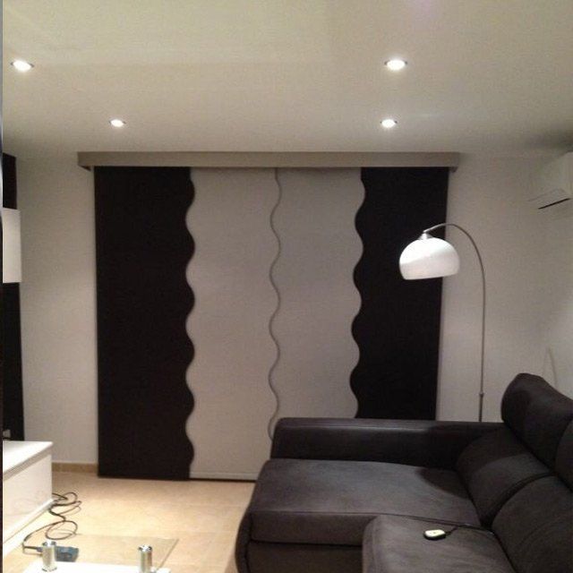 Sala de estar moderna con sofá seccional oscuro, lámpara de pie, luces empotradas y cortinas corredizas en blanco y negro.