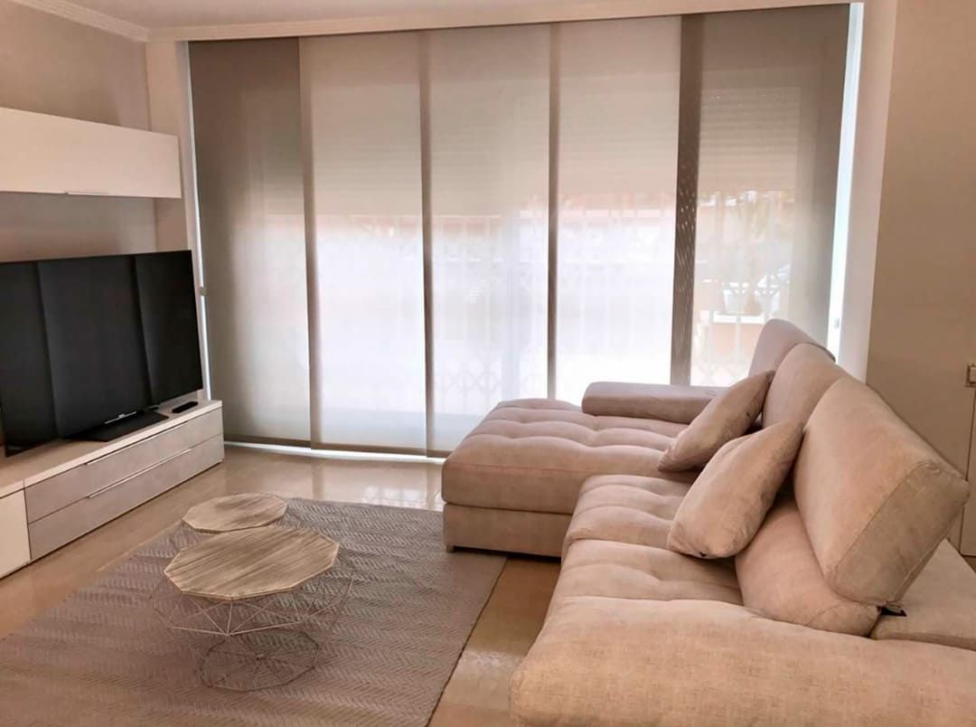 Sala de estar luminosa con sofá modular beige, televisor, alfombra y persianas grandes.