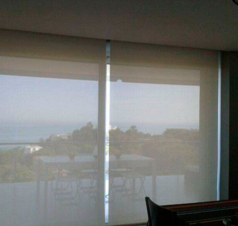 Persianas translúcidas cubren un balcón con vistas al océano y a los árboles al atardecer.