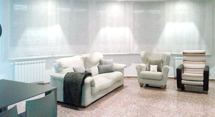 Sala de estar minimalista y luminosa con sofá blanco, sillón, mesa auxiliar y libros apilados bajo una iluminación tenue en la pared.