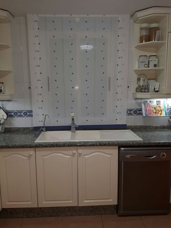 Fregadero de cocina bajo una ventana con cortinas transparentes, armarios beige, encimera de granito y lavavajillas a la derecha.