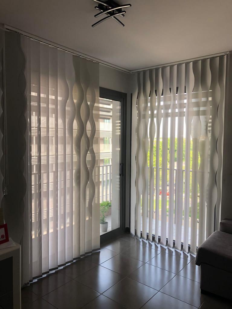 Sala de estar luminosa con cortinas blancas transparentes, grandes ventanales y un sofá gris en la esquina.