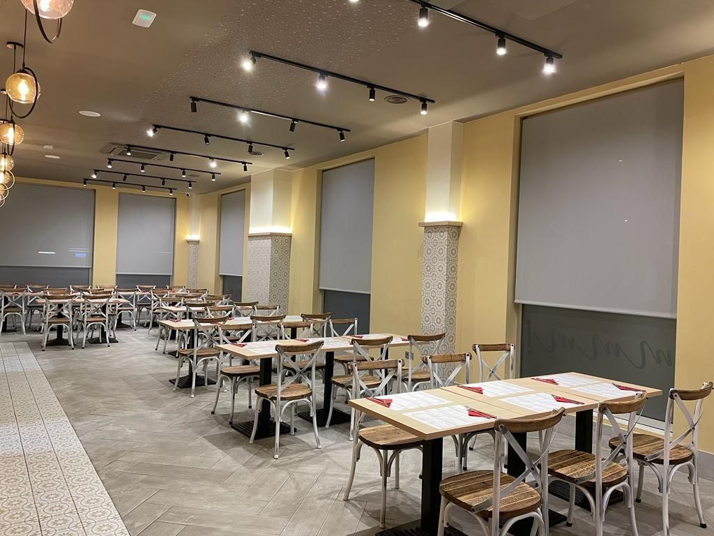 Aula o sala de conferencias vacía, con filas de mesas y sillas, persianas bajadas en las grandes ventanas y focos empotrados en el techo.