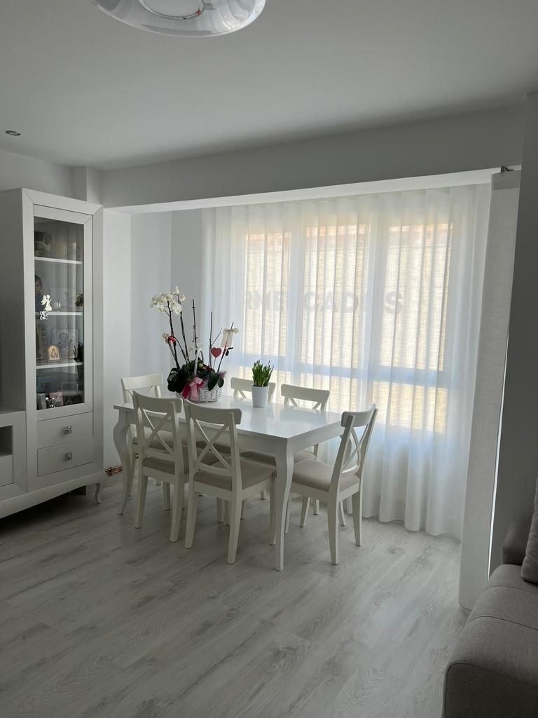 Comedor luminoso con mesa y sillas blancas, flores y cortinas transparentes sobre una ventana soleada.