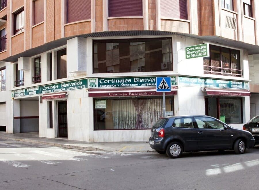 Local comercial en esquina con letrero verde en una calle de la ciudad, con un coche negro aparcado delante.