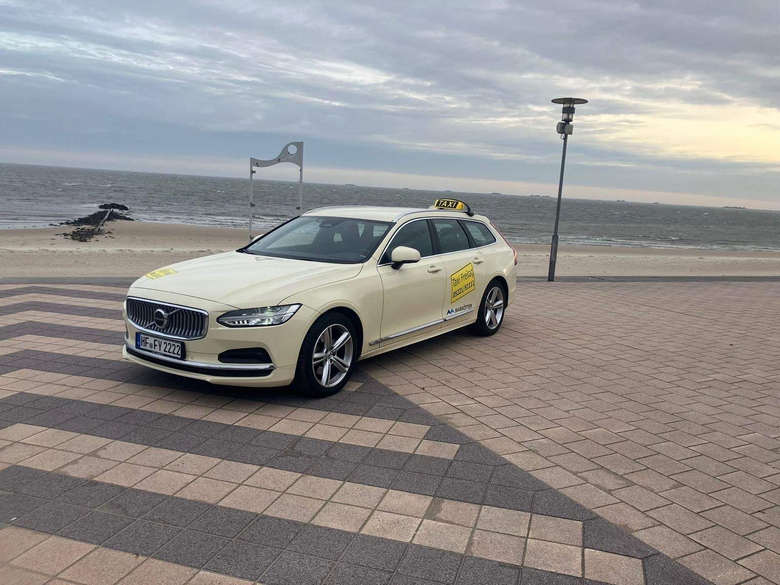 Ein weißes Taxi parkt an einer Strandpromenade, im Hintergrund ist das Meer zu sehen.