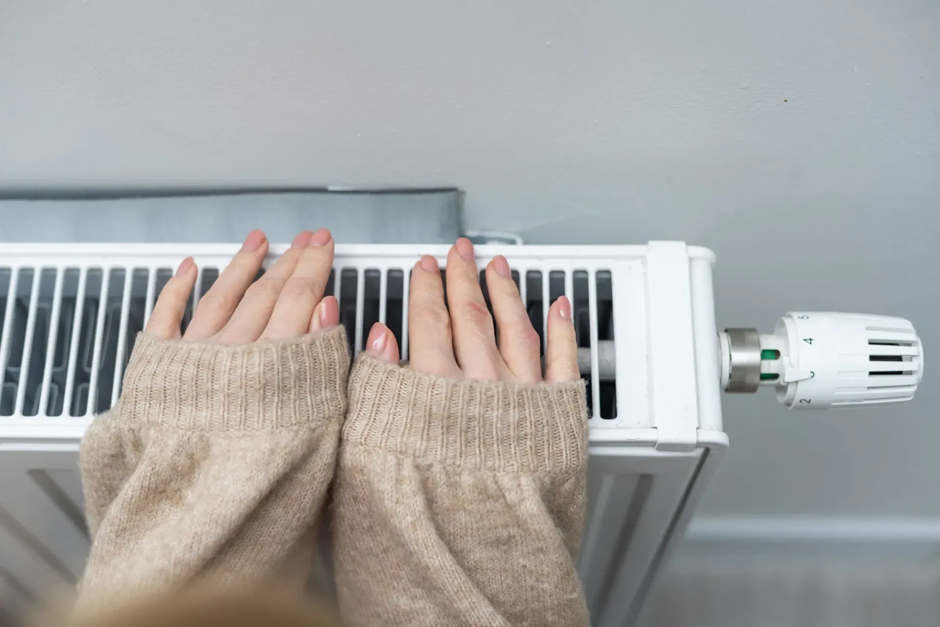 Unas manos, cubiertas con mangas largas de color canela, descansan sobre un radiador blanco