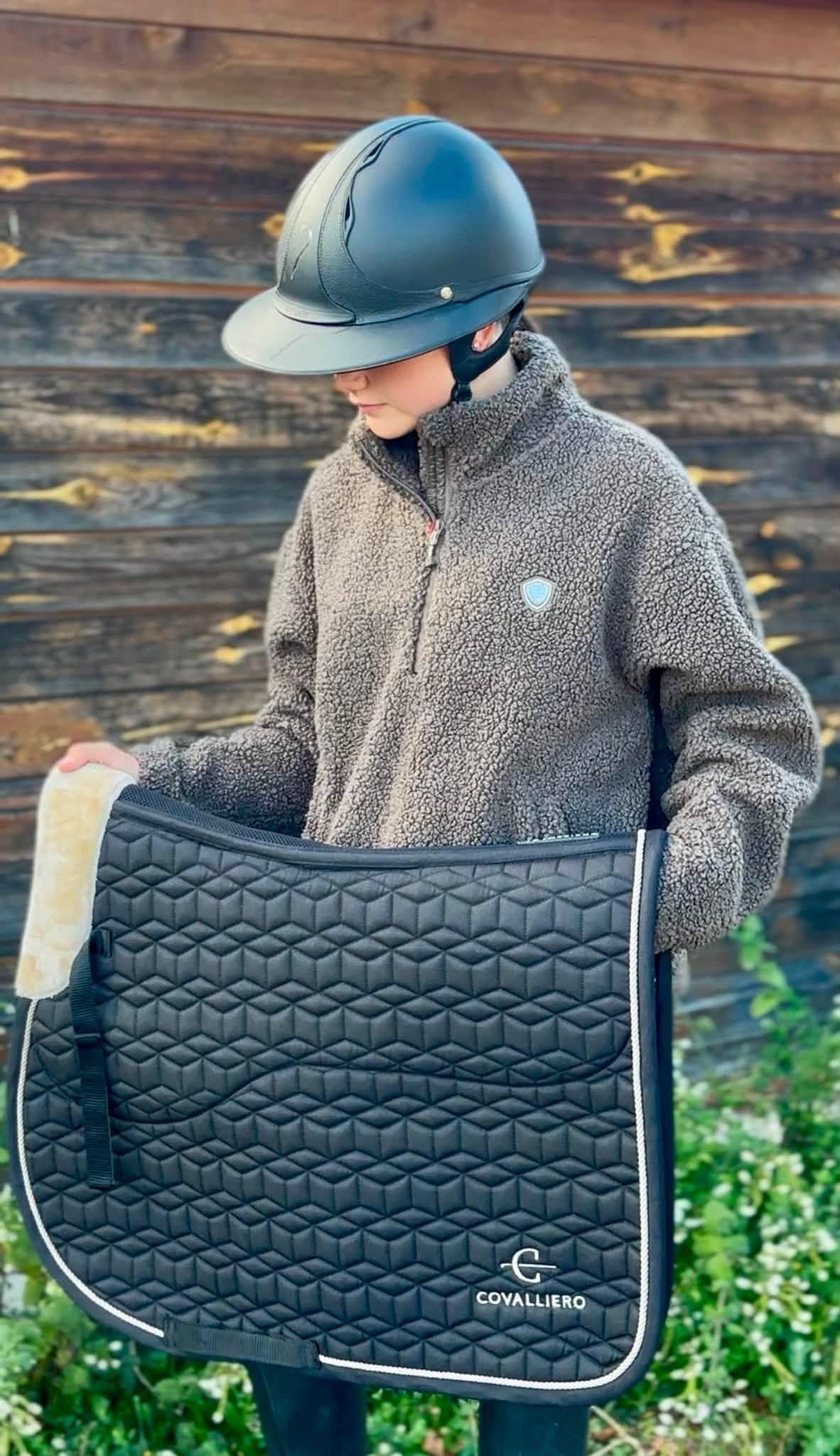 Une personne portant un casque d’équitation tient un tapis de selle noir matelassé devant un mur en bois.