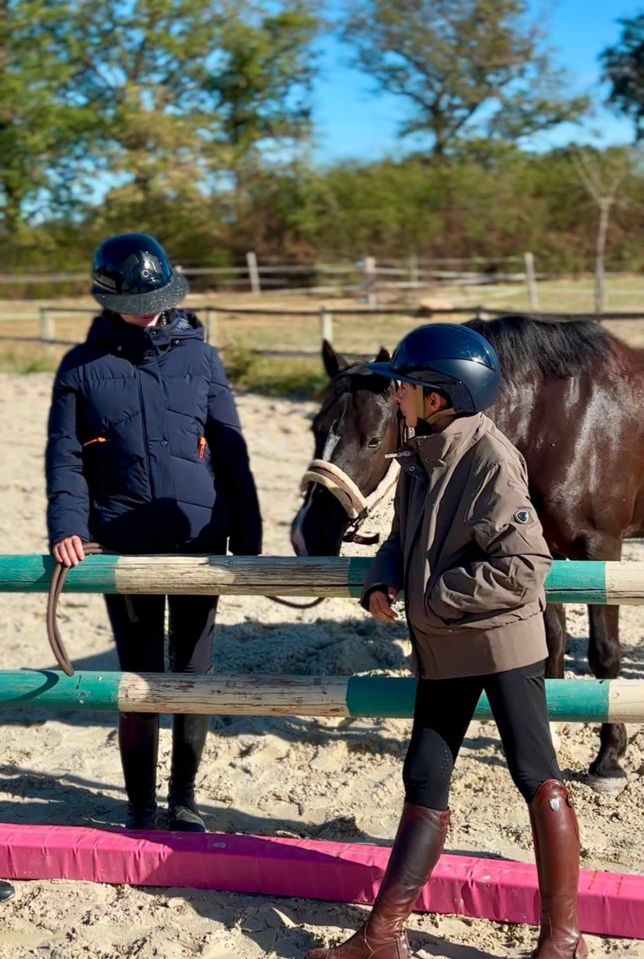 Deux cavaliers portant un casque se tiennent à côté d’un cheval dans un manège en plein air ensoleillé
