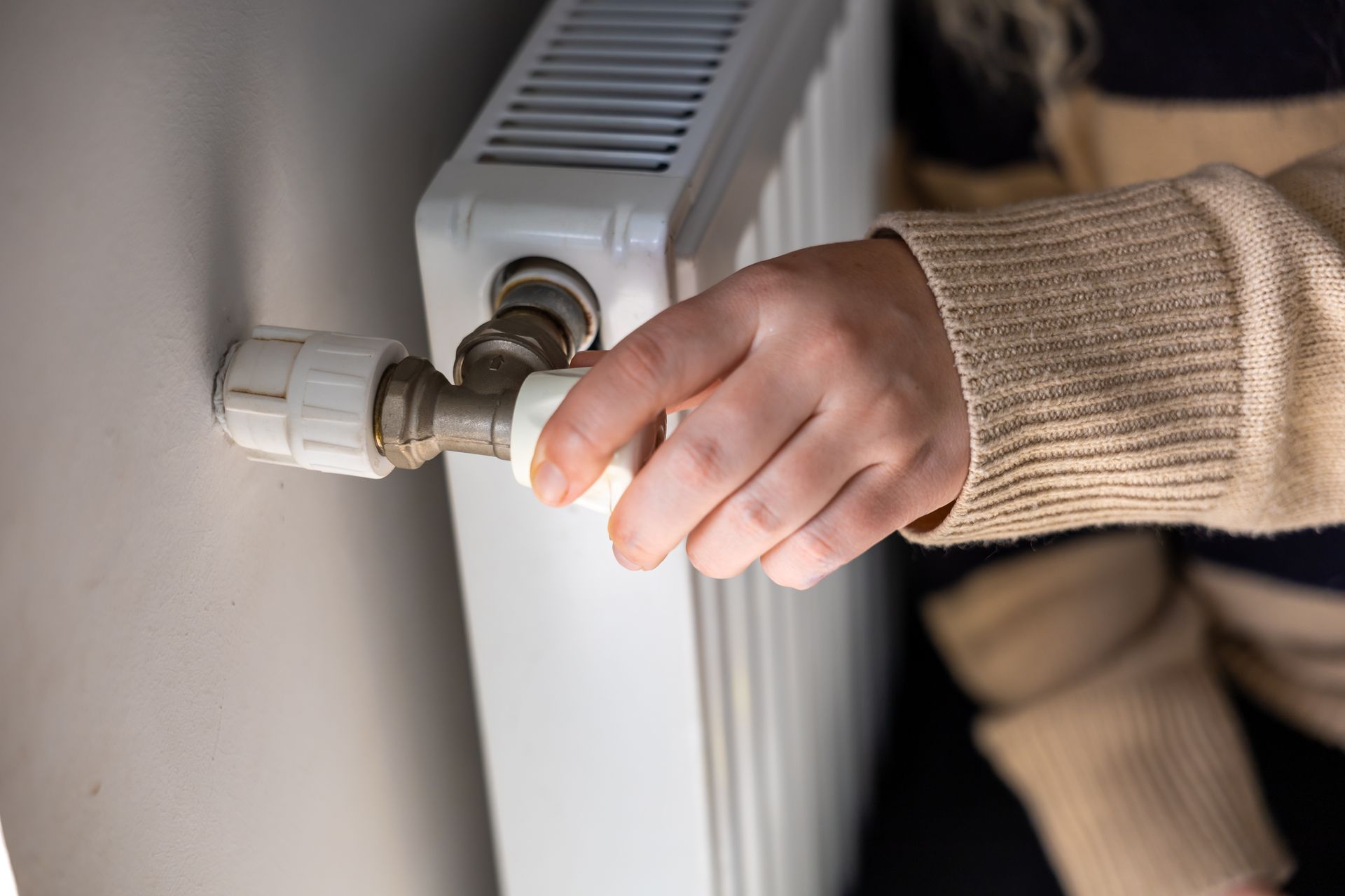 Une main actionne un robinet de radiateur à côté d'un radiateur blanc, laissant apparaître la manche d'un pull beige.