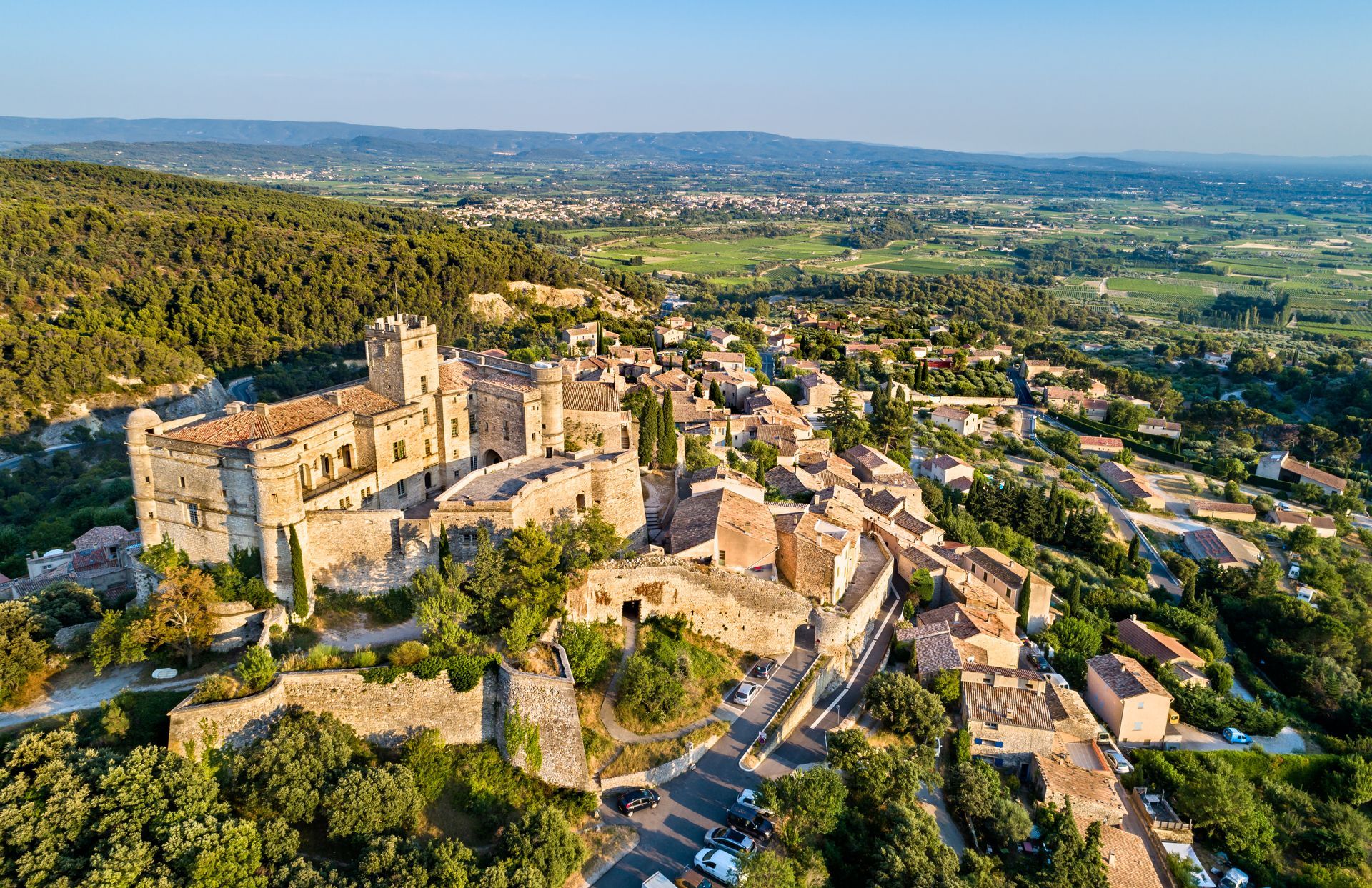 Vue aérienne d'un village de pierre à flanc de colline, avec les ruines d'un château et la campagne verdoyante s'étendant à perte de vue.