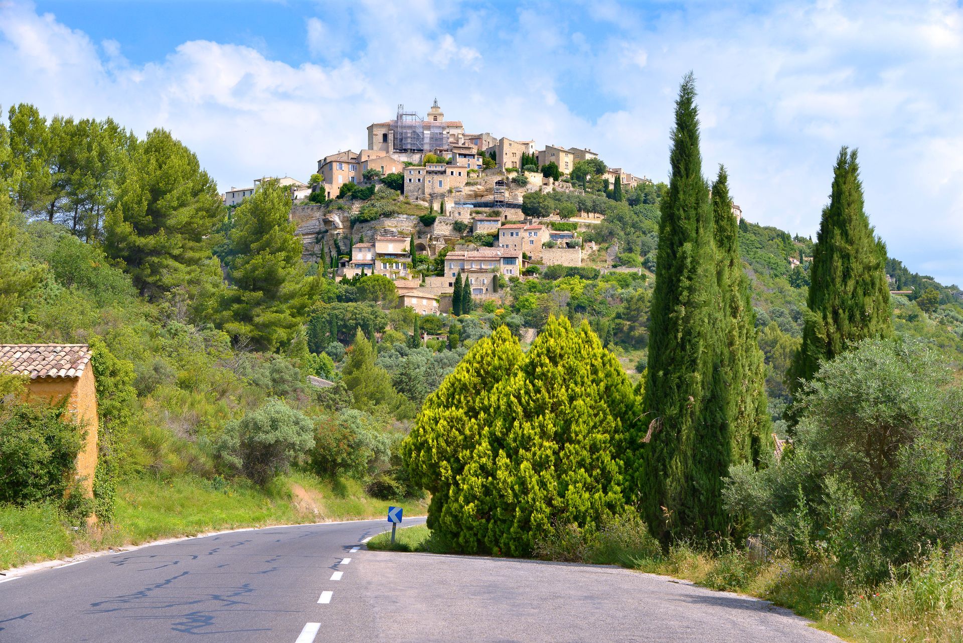 Route sinueuse menant à un village perché sur une colline, sous un ciel bleu, encadrée d'arbres verts et de cyprès.