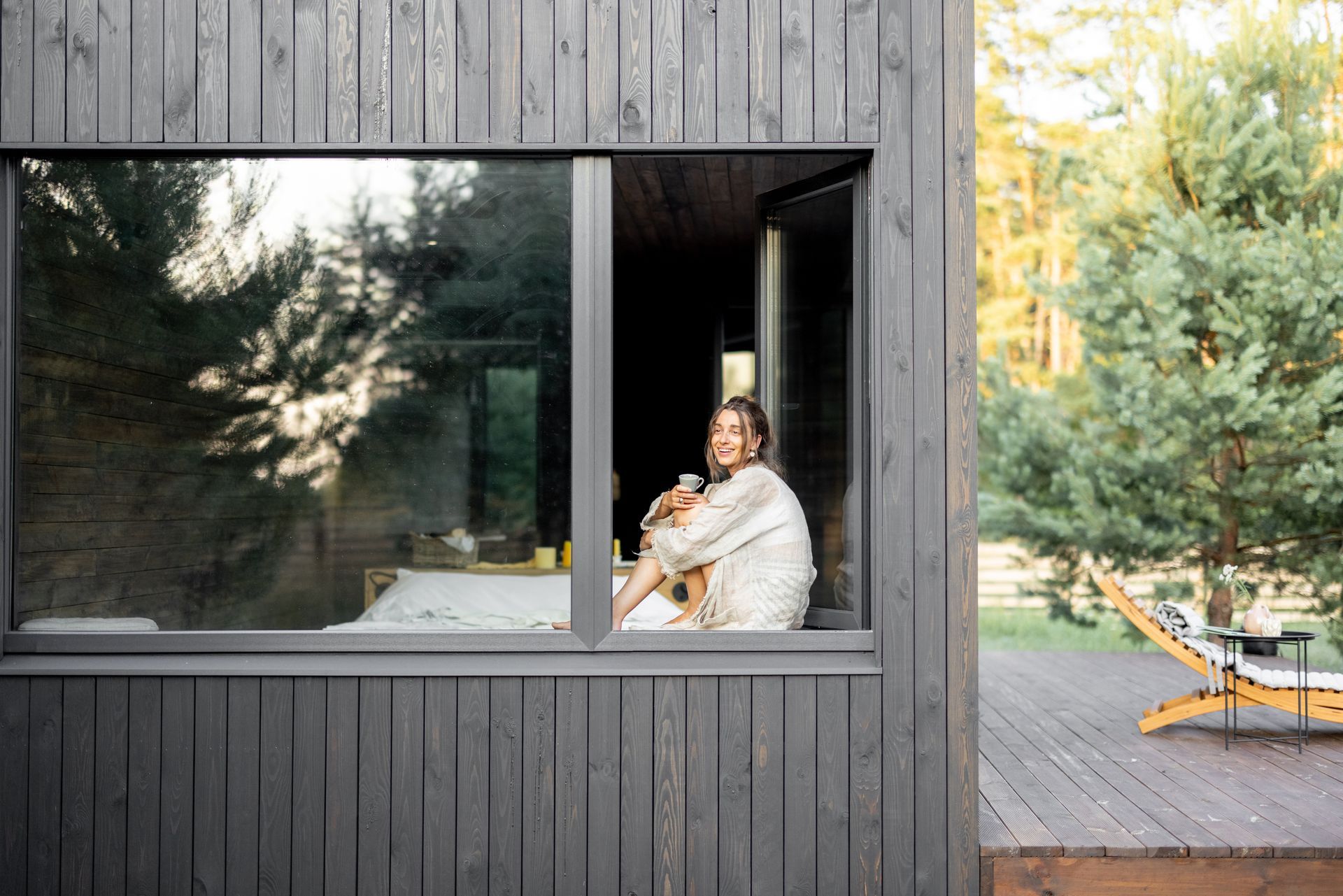 Une personne assise près d'une grande fenêtre dans une cabane en bois sombre, donnant sur une terrasse boisée et des arbres.