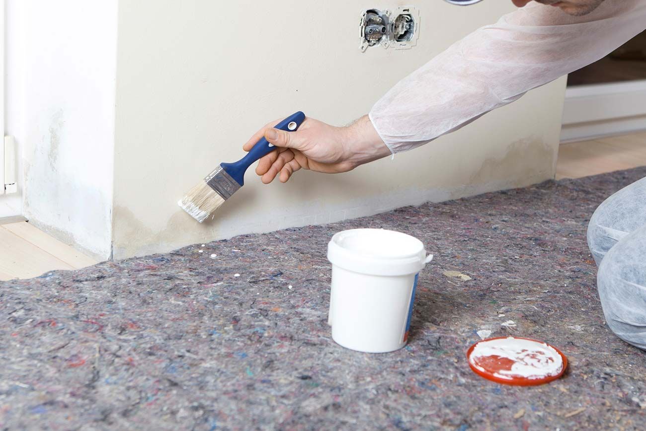 Persona pintando una pared blanca con un pincel, lata de pintura en el suelo, revestimiento de suelo gris.