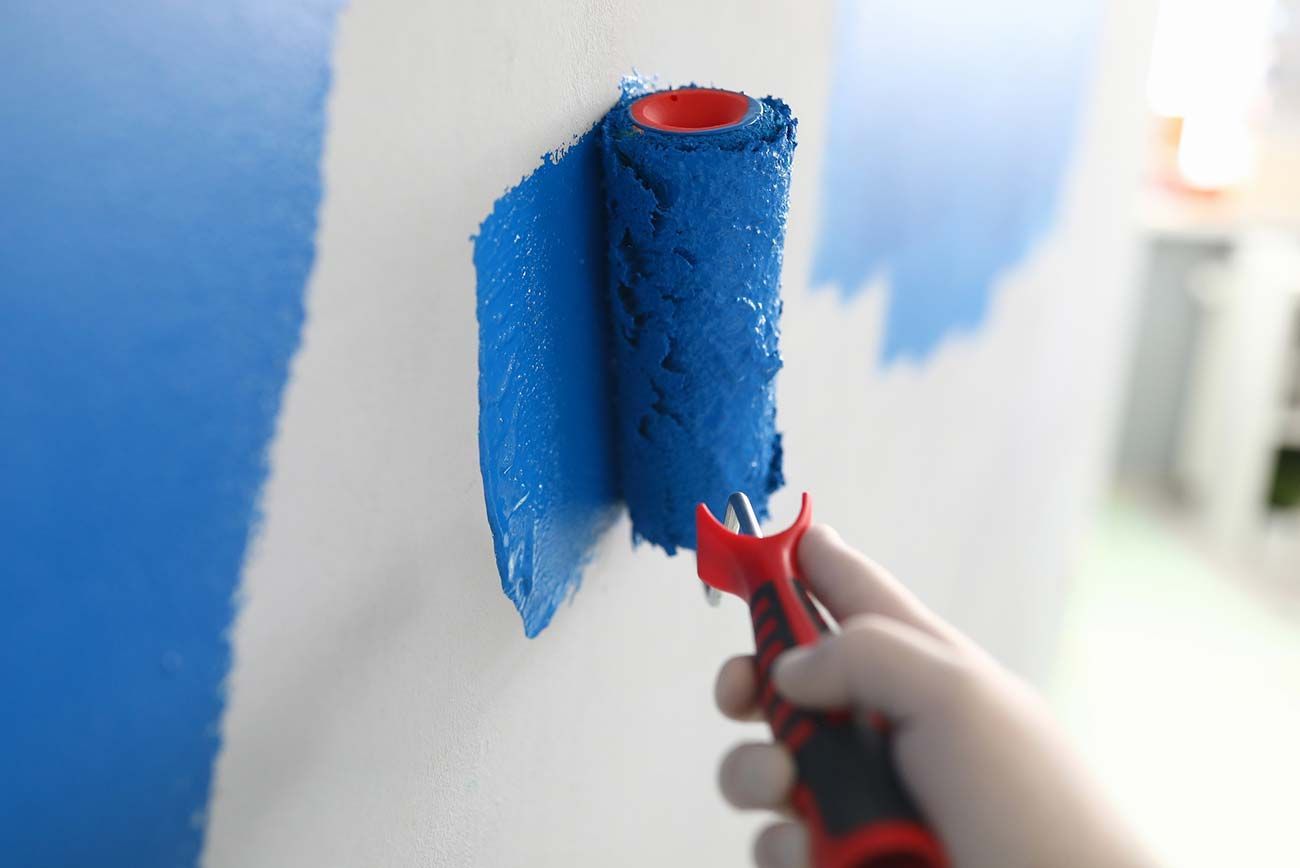Mano sosteniendo un rodillo de pintura, aplicando pintura azul sobre una pared blanca.