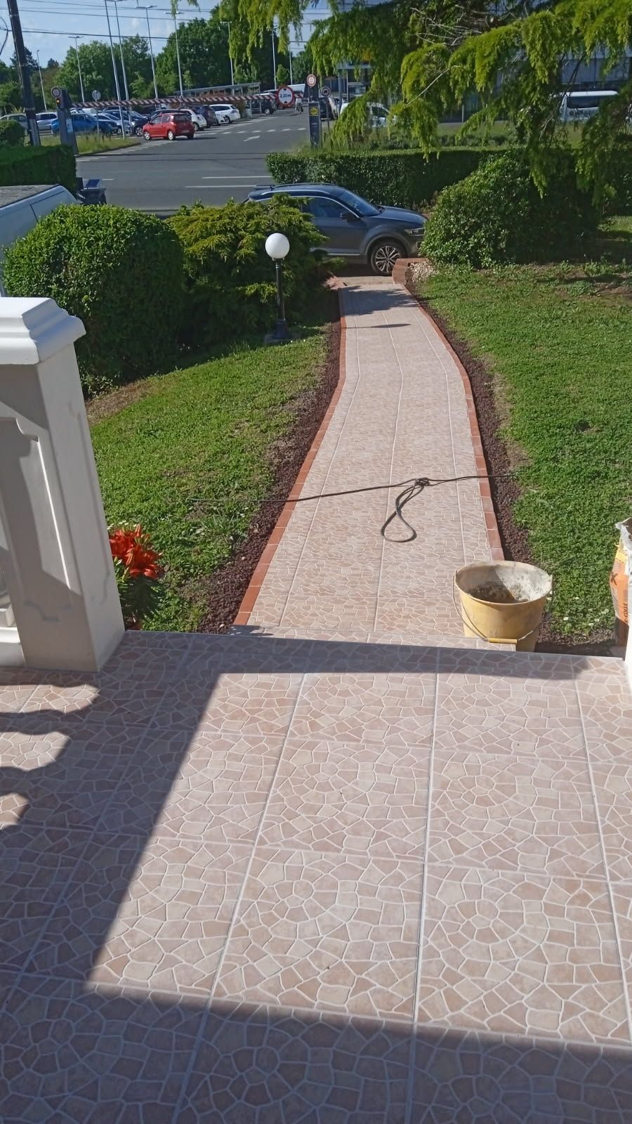 Une allée carrelée menant à la rue, bordée d'herbe et de buissons ; un porche d'immeuble se trouve au premier plan.