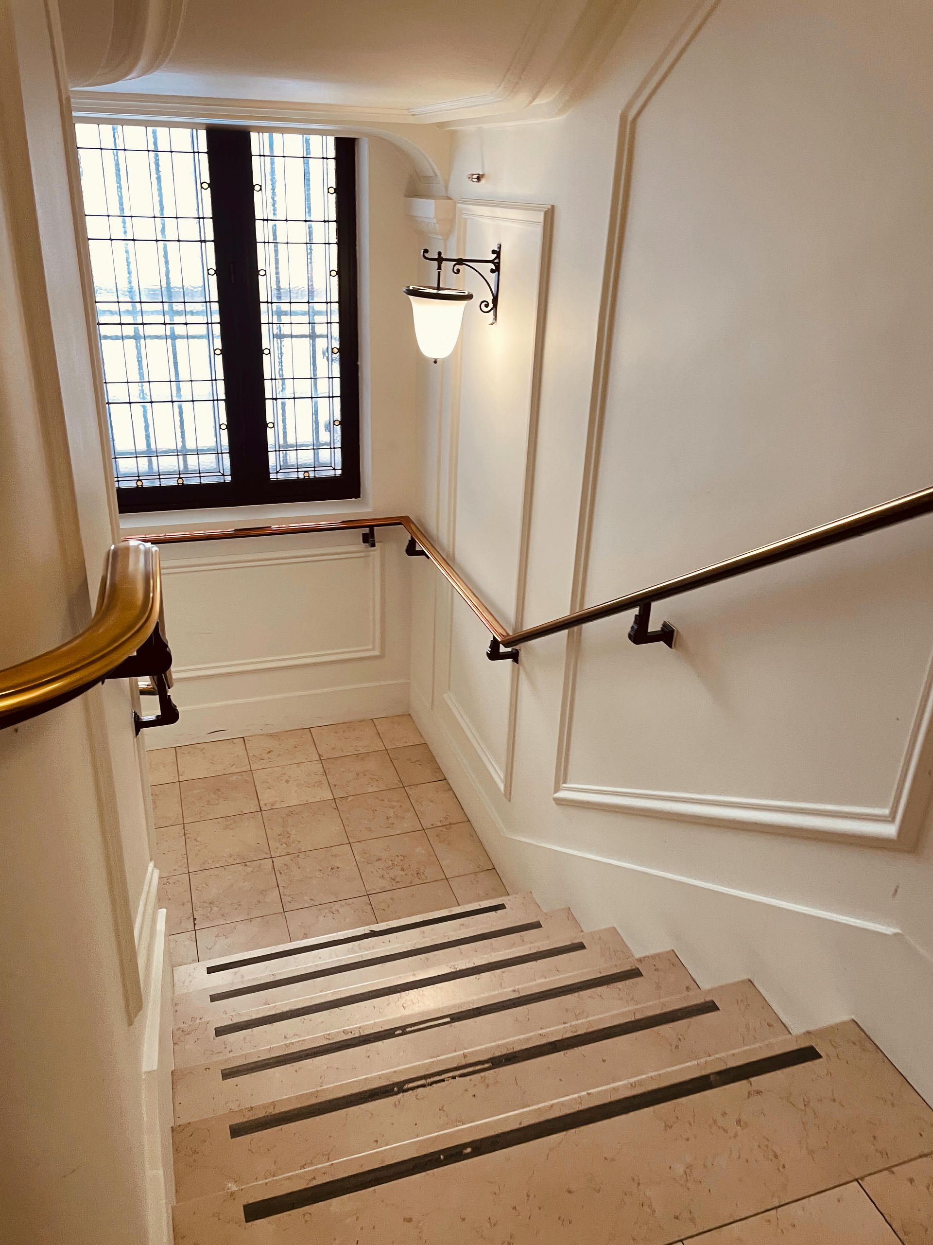 Escalier avec marches beiges, rampes et fenêtre. Les murs sont blancs et éclairés par une applique.