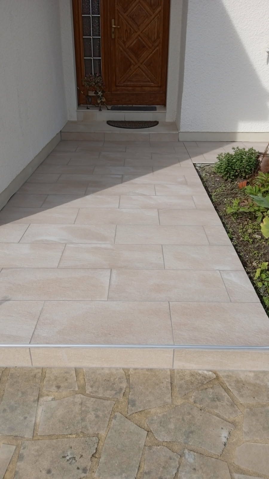 Chemin carrelé beige menant à une porte en bois marron, avec un petit parterre de jardin à droite.
