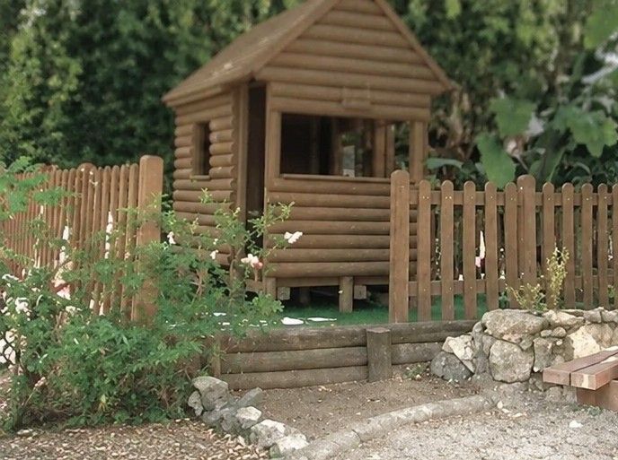Pequeña cabaña de madera en un jardín, detrás de una valla de listones, con rocas y vegetación cerca.