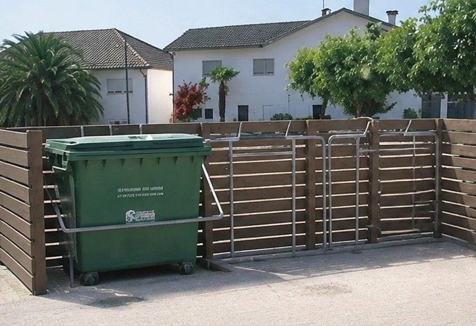Contenedor verde de reciclaje junto a una valla de listones marrones en la entrada de una casa de las afueras, con casas al fondo