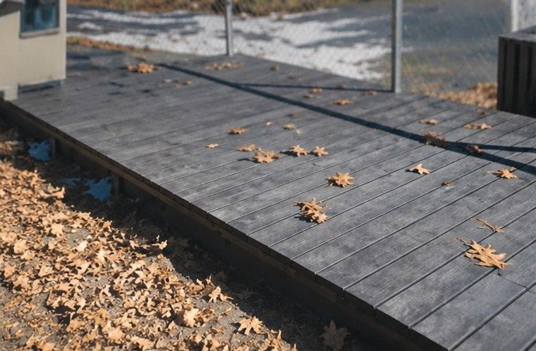 Tarima de madera cubierta de hojas marrones caídas, junto a una barandilla y un pavimento exterior