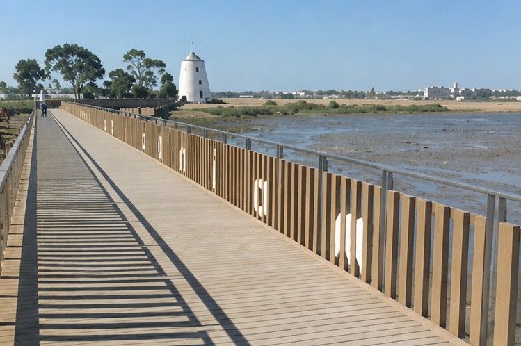 Pasarela de madera junto a un río, con una barandilla y un faro a lo lejos bajo un cielo despejado