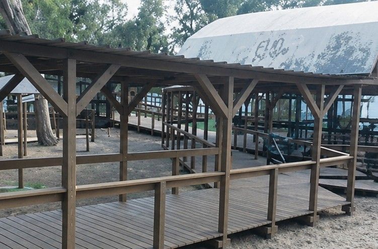 Pabellón de madera con barandillas y techo de chapa ondulada en un terreno de tierra entre árboles