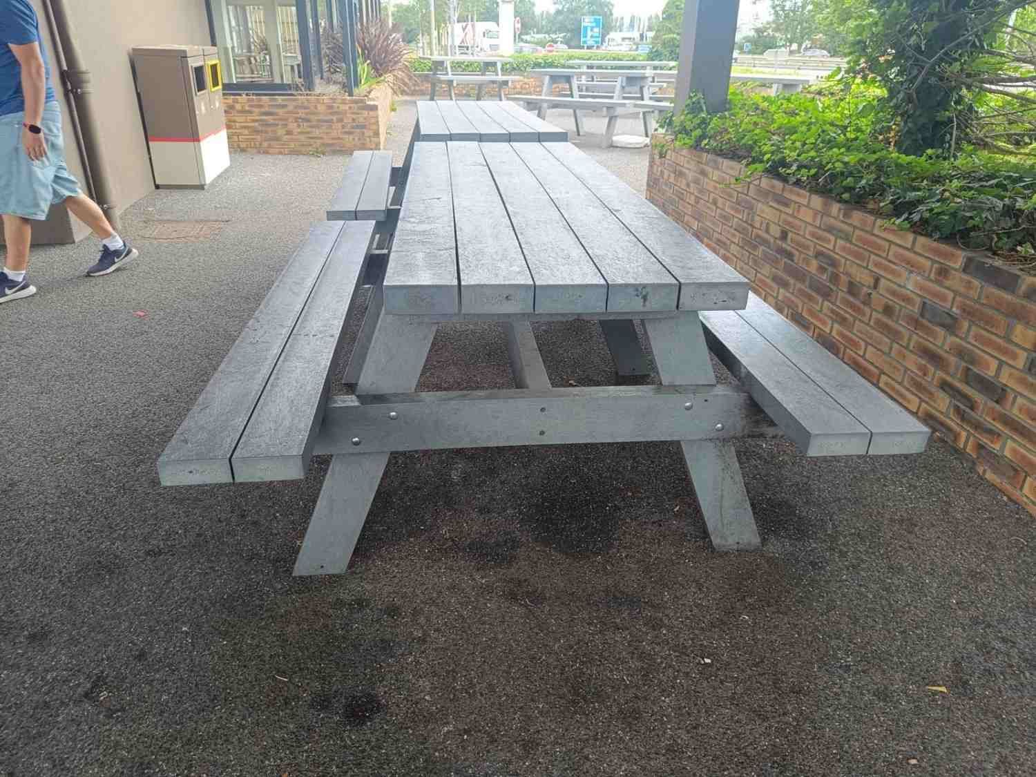 Mesa de picnic gris en un patio de grava junto a una pared de ladrillo, con una persona pasando por la izquierda.