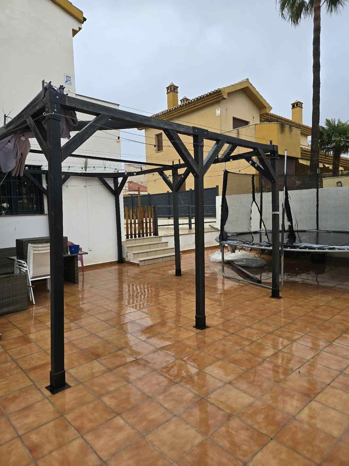 Patio embaldosado empapado por la lluvia, con estructuras negras de pérgola y muebles de exterior, junto a unas casas y palmeras