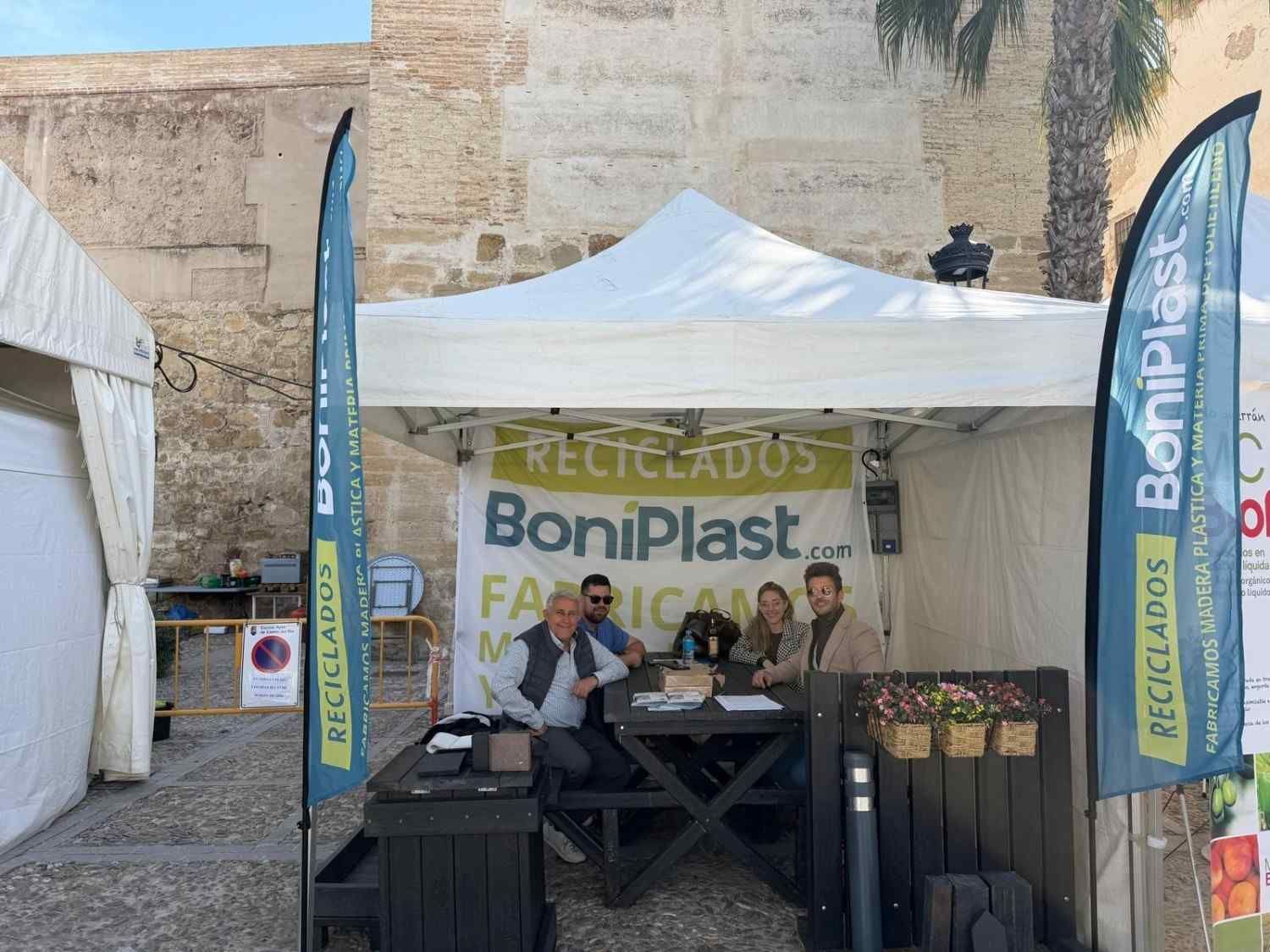 Puesto de la feria BonsaiPlast al aire libre bajo una carpa blanca, con dos personas detrás de una mesa de exhibición y varias pancartas.