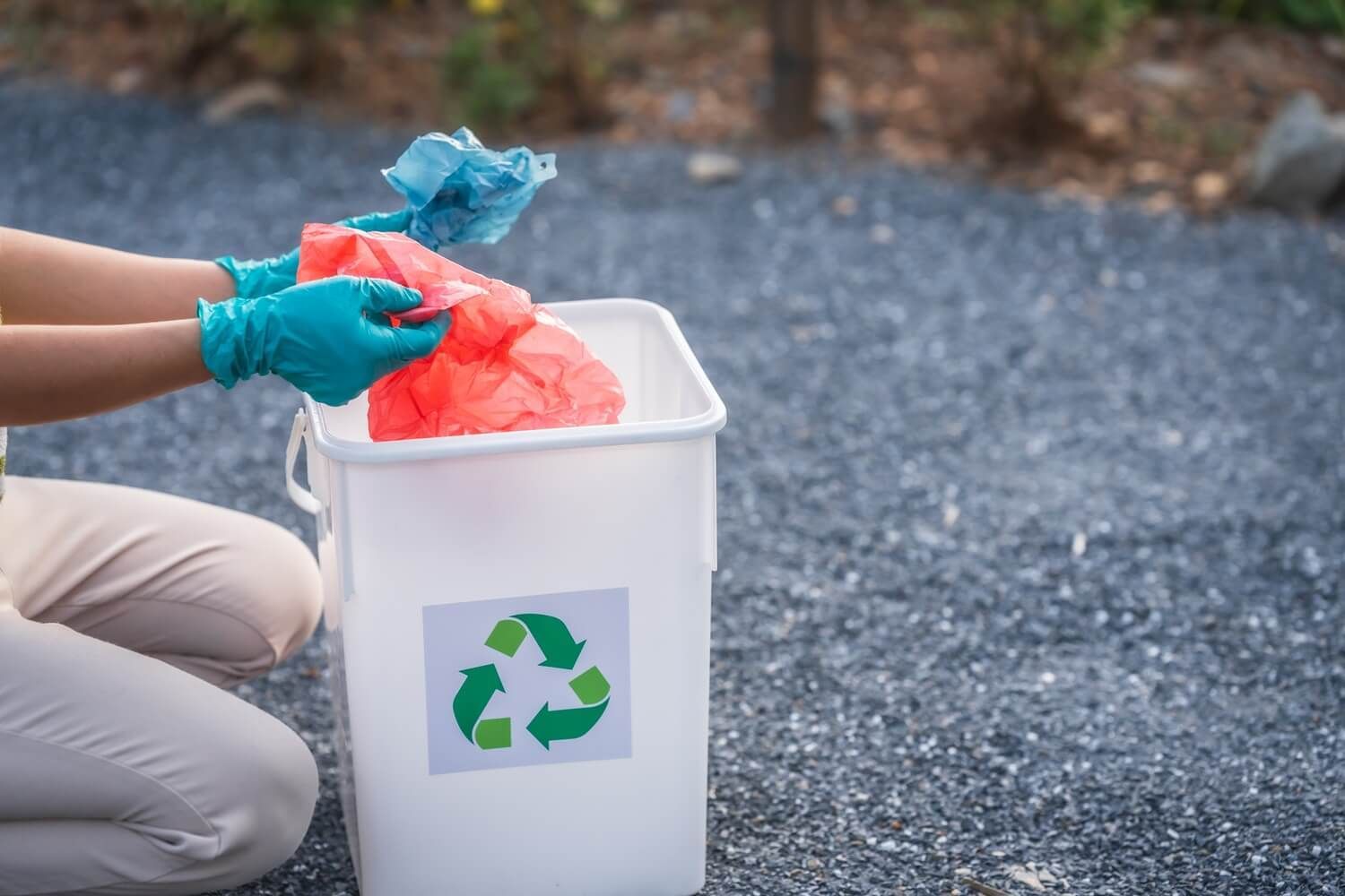 Manos depositando botellas de plástico en un contenedor de reciclaje sobre grava al aire libre