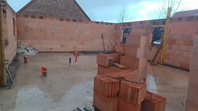 Chantier de construction avec des murs en briques, un sol en béton avec des tuyaux de plomberie et une pile de briques.