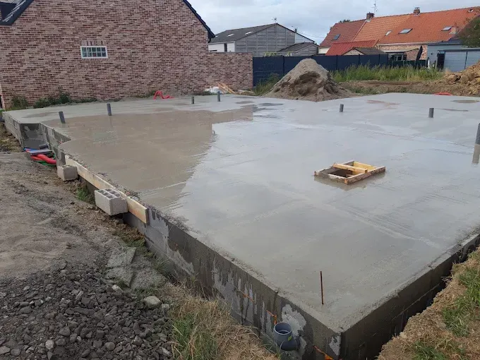 Fondations en béton fraîchement coulées pour un projet de construction, situées dans une cour résidentielle avec des maisons en arrière-plan.
