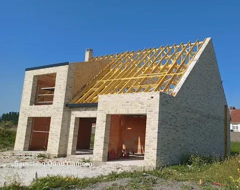 Une maison à deux étages partiellement construite, avec des murs en pierre et une charpente de toit en bois apparente.