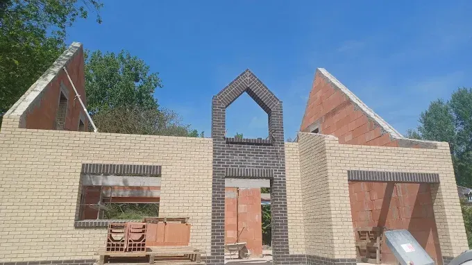 Une maison en construction avec des murs en briques, une entrée voûtée décorative sombre et des pignons en briques apparentes.