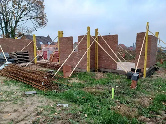 Des murs de briques en construction sur un terrain herbeux, soutenus par des poutres de contreventement en bois, se dressent sous un ciel nuageux.