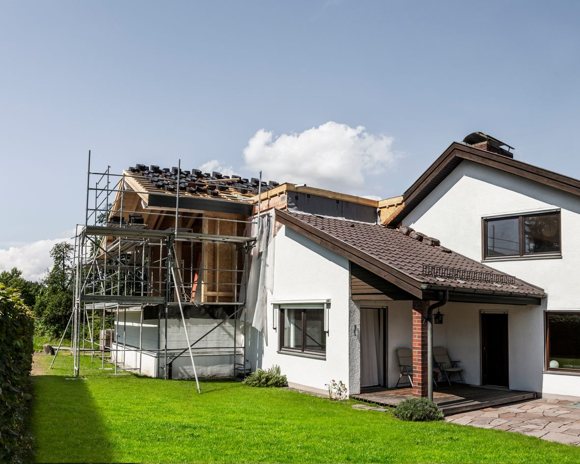 Une maison à deux étages, au toit partiellement rénové et soutenu par des échafaudages, par une journée ensoleillée, avec une pelouse.