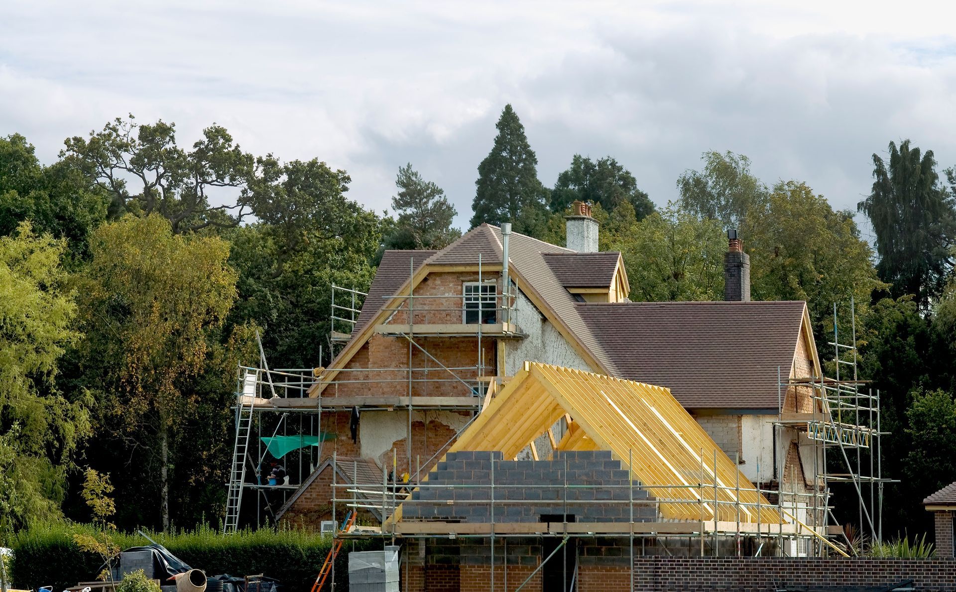 Une maison à plusieurs étages en construction, avec des fermes de toit en bois apparentes et des échafaudages, entourée d'arbres.