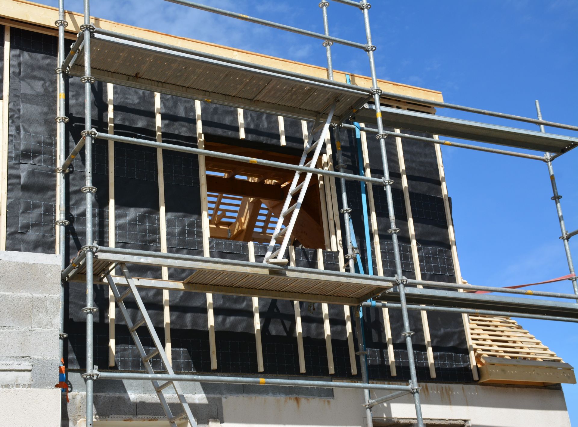 Des échafaudages entourent l'extérieur inachevé d'un bâtiment dont la charpente en bois et la membrane d'étanchéité sont visibles.