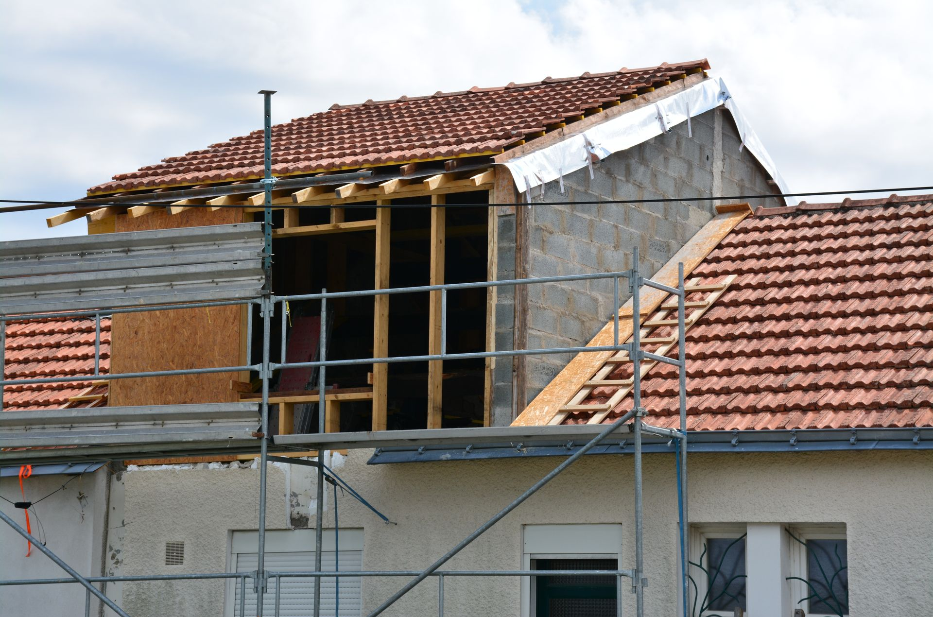 Une maison en construction avec des échafaudages, présentant une nouvelle structure en bois au deuxième étage, encore inachevée, et un toit de tuiles.