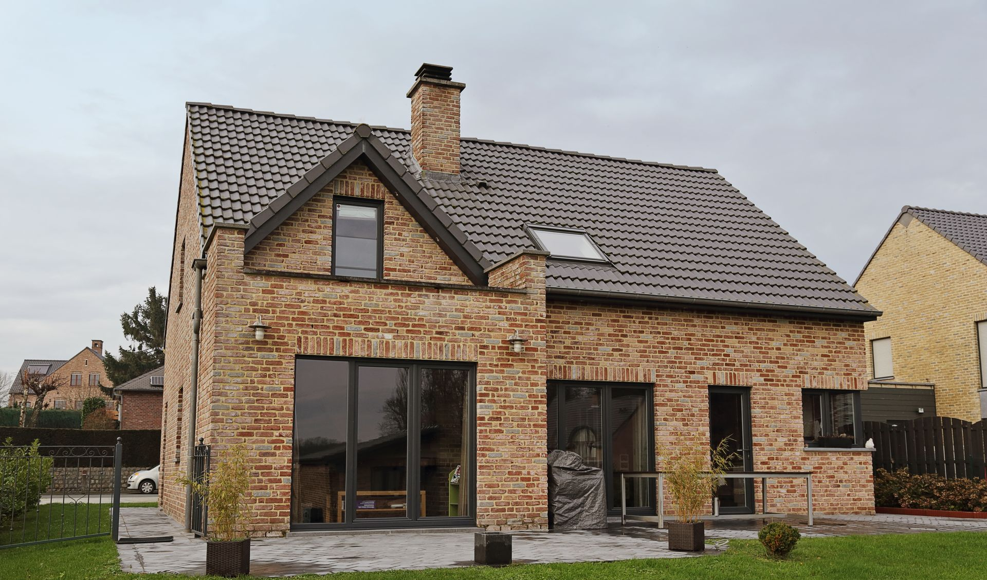 Une maison à deux étages avec une façade en briques, un toit de tuiles foncées et de grandes portes vitrées donnant sur un patio.