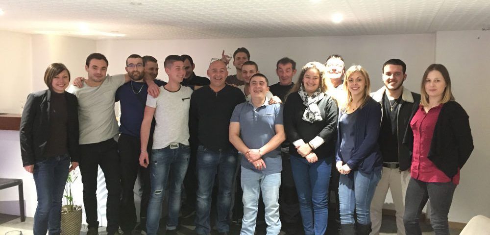 Un groupe d'une vingtaine de personnes, pour la plupart des jeunes adultes, se tiennent serrées les unes contre les autres à l'intérieur, souriant à la caméra.