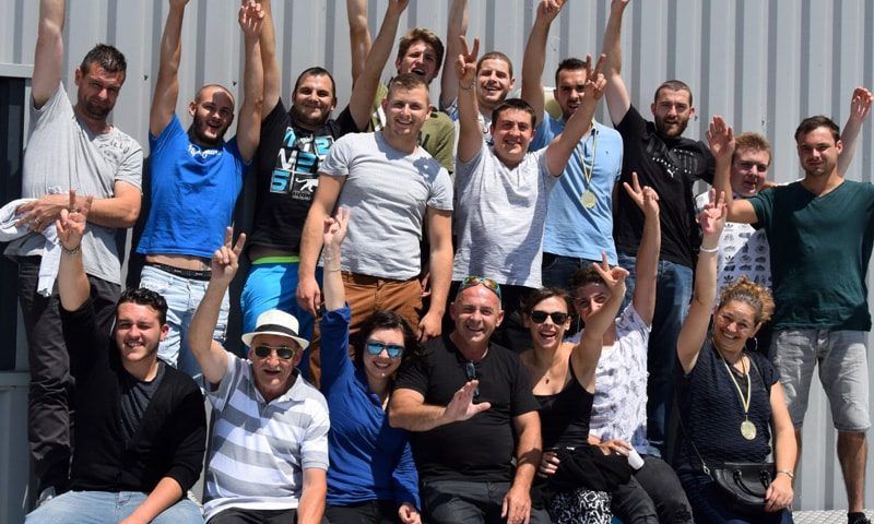Groupe de personnes souriantes et levant les bras devant un mur blanc ; certaines brandissent des signes de paix.