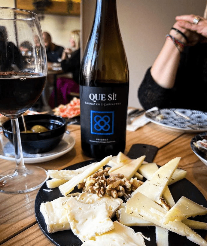 Una botella de que se encuentra en una mesa al lado de un plato de queso y nueces.
