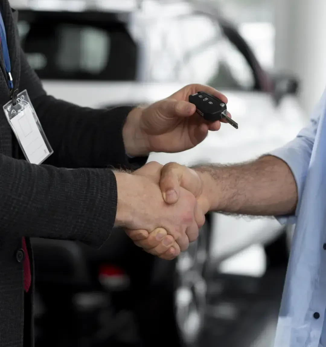Persona con traje entregando las llaves de un coche a otra persona, estrechándole la mano en un concesionario de automóviles.