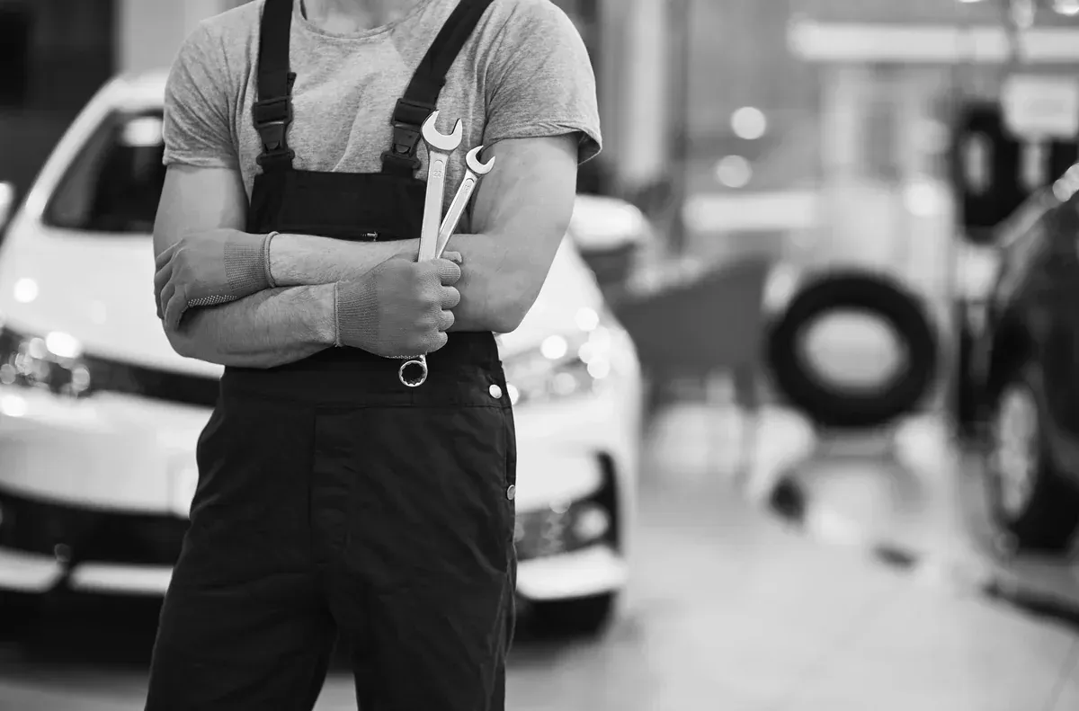 Mecánico con mono sosteniendo llaves inglesas en un taller de reparación de automóviles, con automóviles y neumáticos en el fondo.