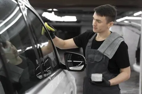 Un hombre con un mono limpia la ventana de un coche con un paño amarillo, en un garaje.