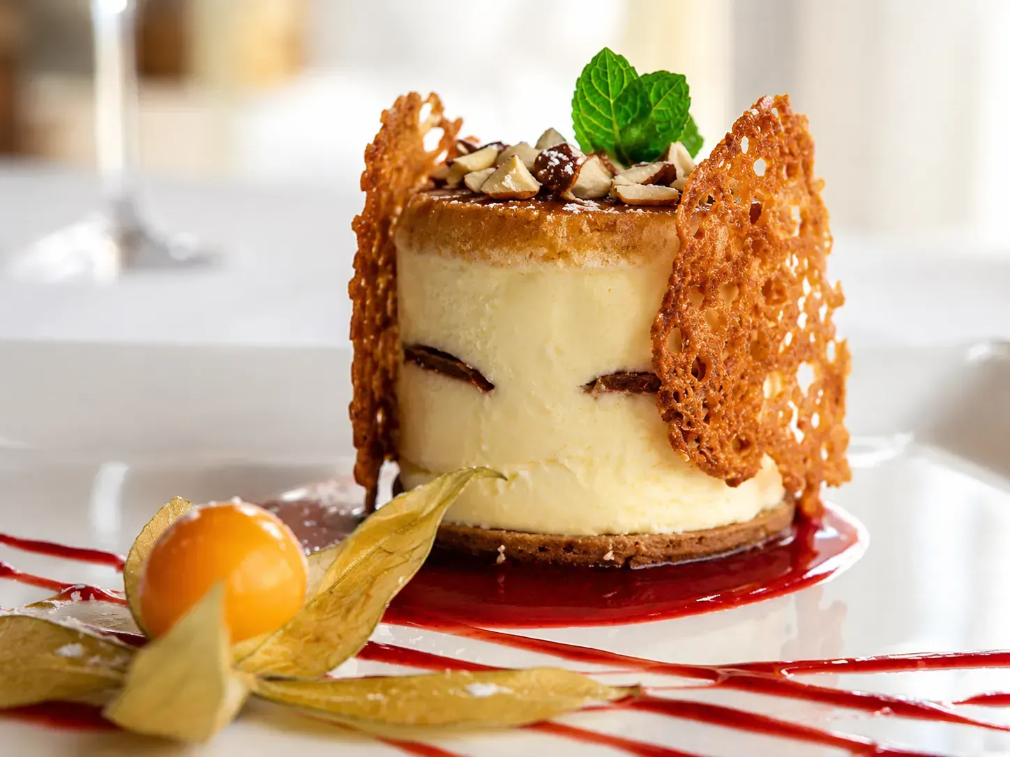 Pastel de postre emplatado con guarnición de caramelo, hoja de menta y salsa roja sobre una mesa blanca.